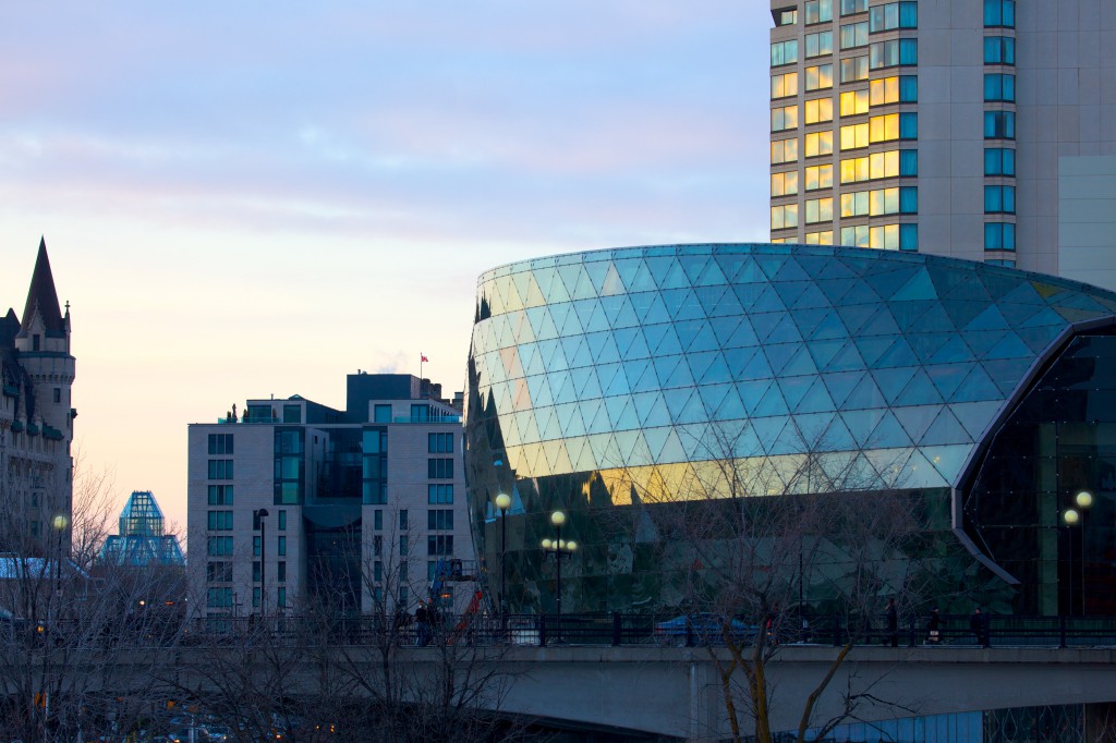 An evening view of the Ottawa Convention Centre (a.k.a. The Shaw Centre) taken from south of the building looking north. The photo shows the bowing exterior glazed wall of the 4 storey OCC. Adjacent to the OCC is the Westin Hotel, with the tower of the historic Frontenac Chateau Laurier also visible on the far left of the photo.